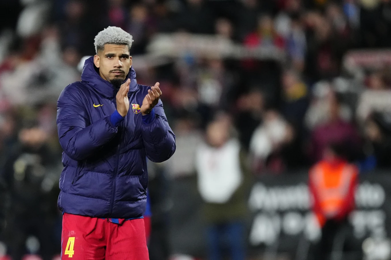 Piłkarz ubrany w klubową kurtkę i czerwone spodenki, o jasnych włosach, stoi na tle rozmytego tłumu i bije brawo.