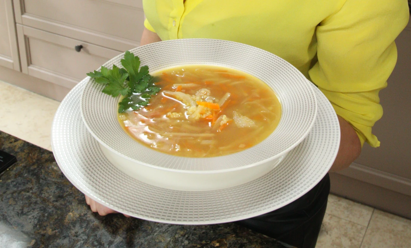 Talerz gorącej zupy z warzywami i natką pietruszki na wierzchu trzymany przez osobę ubraną w żółtą bluzkę, na tle kuchennych szafek.