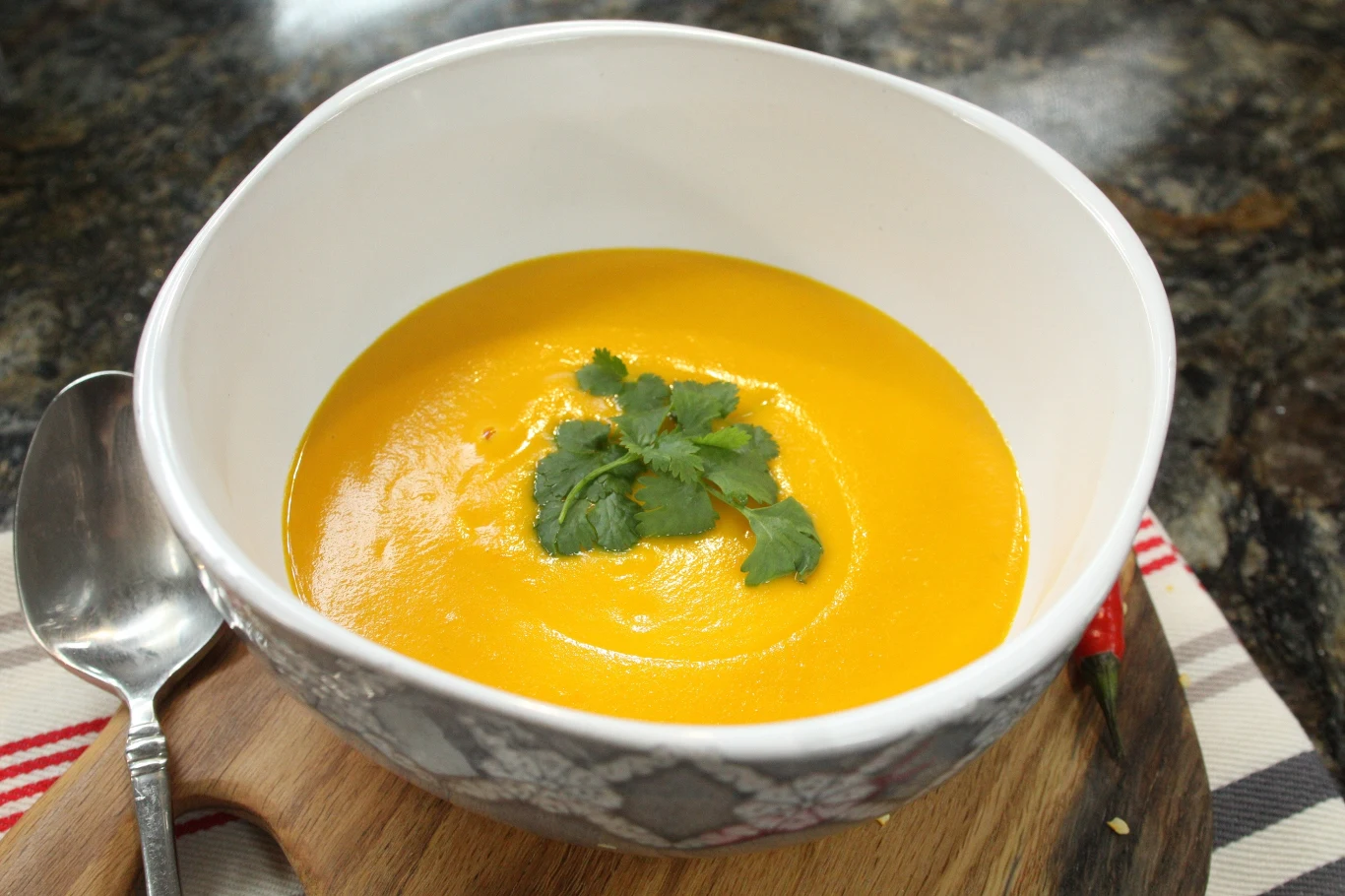 Gładki krem z marchewki w białej misce, ozdobiony świeżą natką pietruszki, położony na drewnianej desce obok łyżki i ścierki w biało-czerwone paski.