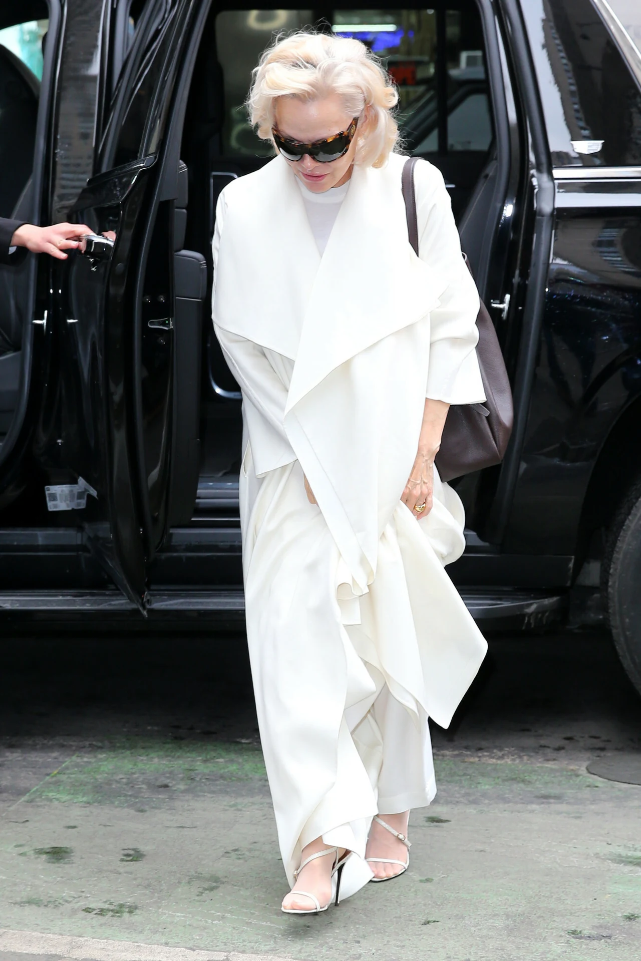 Starsza kobieta ubrana w elegancki, biały strój oraz okulary przeciwsłoneczne wychodzi z luksusowego samochodu, trzymając torebkę na ramieniu.