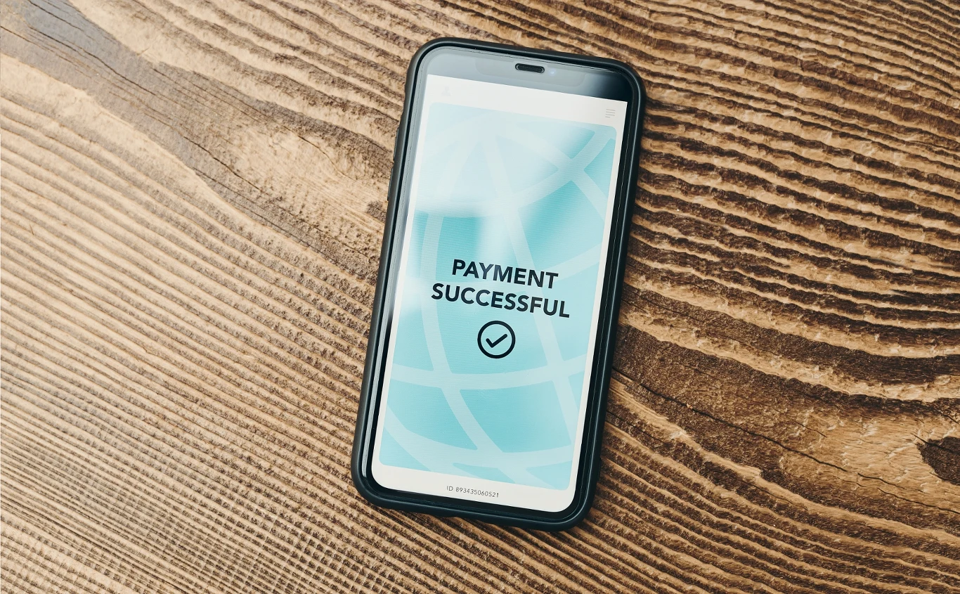 Odnawiające się automatycznie subskrypcje to ogromne koszty Smartfon leżący na drewnianym stole, na ekranie widoczny komunikat o pomyślnie zakończonej płatności, tło ekranu w odcieniach niebieskiego, obudowa telefonu czarna.