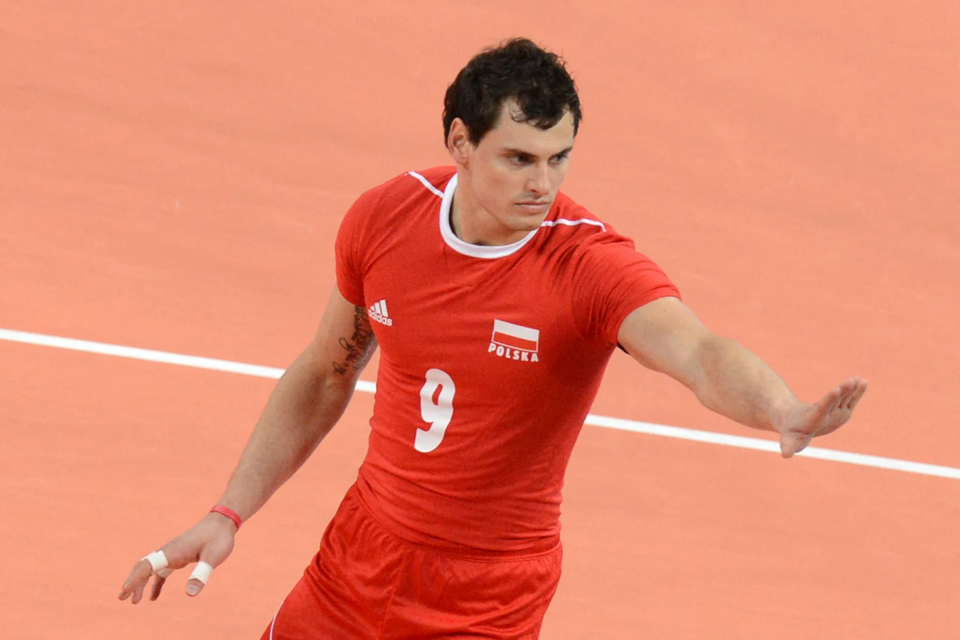 Siatkarz w czerwonym stroju z numerem 9 wykonuje gest ręką na boisku do siatkówki, w tle widać linię boiska.