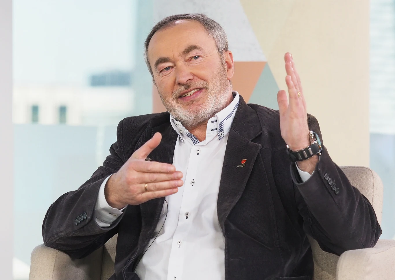 Mężczyzna w średnim wieku siedzący na jasnym fotelu, ubrany w białą koszulę i ciemną marynarkę, wykonuje gesty rękami podczas rozmowy i uśmiecha się, w tle jasne, rozmyte wnętrze.