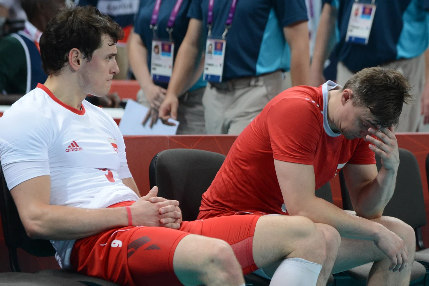 Dwóch siatkarzy w biało-czerwonych strojach siedzi na ławce, jeden z nich trzyma głowę w dłoniach w geście rozczarowania, drugi patrzy przed siebie z poważnym wyrazem twarzy, w tle osoby zaangażowane w wydarzenie sportowe.