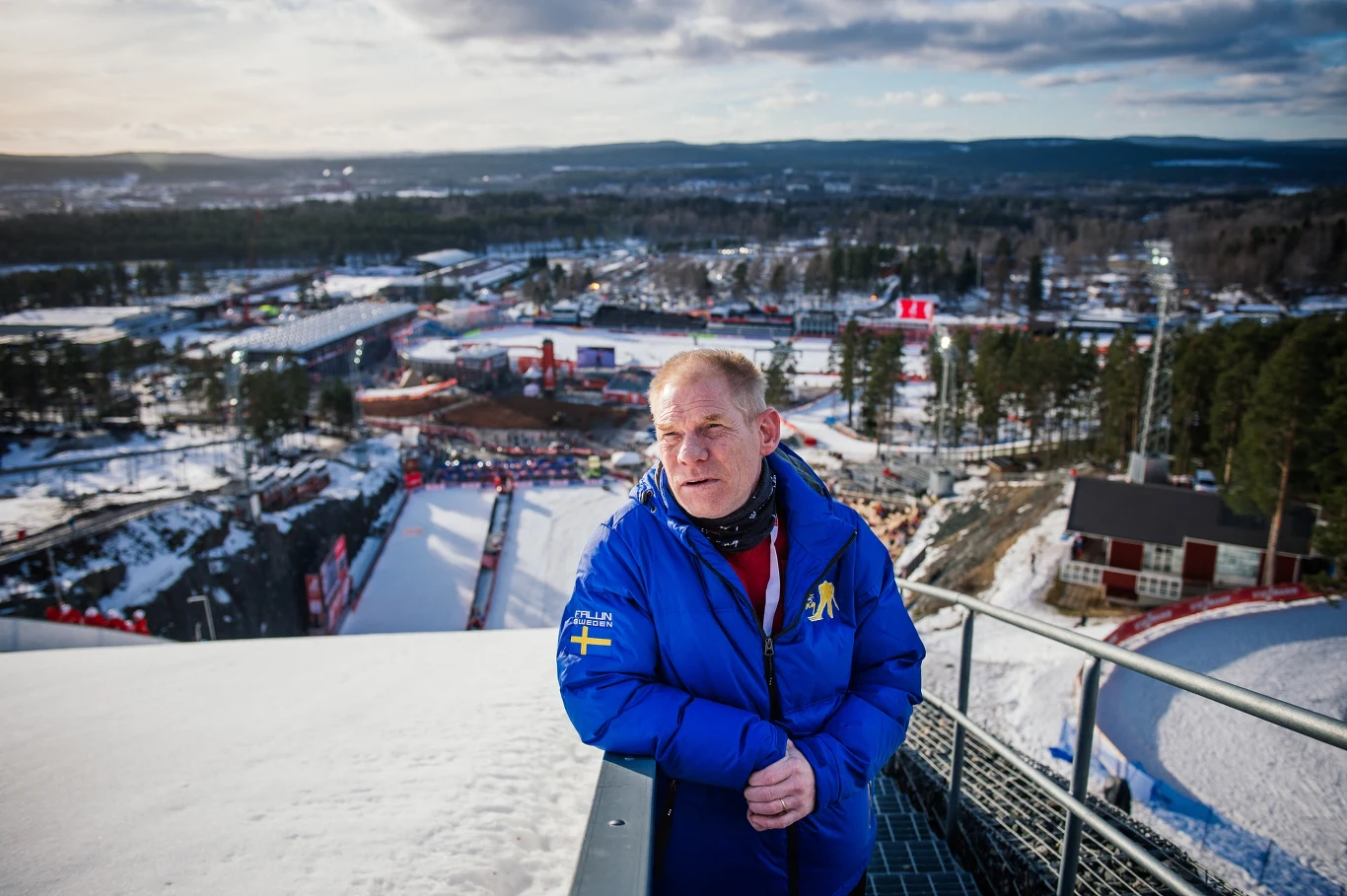 Mężczyzna w niebieskiej kurtce z emblematem Szwecji stoi na tle zimowego krajobrazu, ze skocznią narciarską oraz widocznym w oddali ośnieżonym stadionem sportowym.