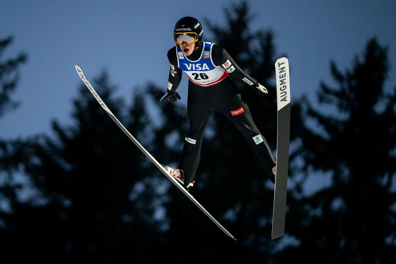 Zawodnik w kombinezonie narciarskim w trakcie lotu podczas skoku narciarskiego na tle wieczornego nieba i sylwetek drzew, widoczny numer startowy oraz logo sponsora na stroju.
