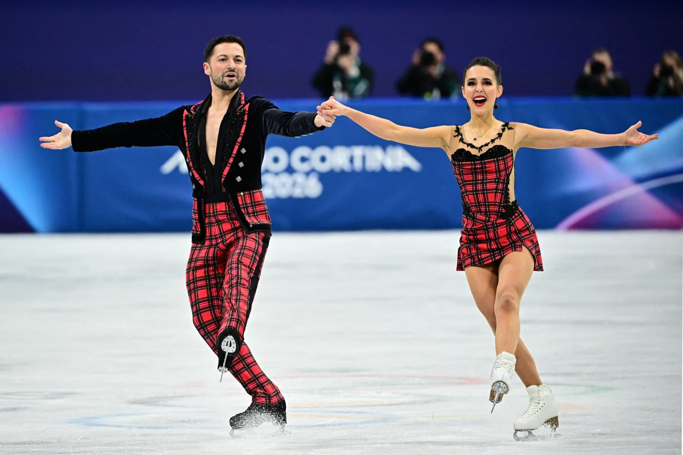 Para łyżwiarzy figurowych wykonujących program na lodowisku, oboje ubrani w charakterystyczne, czerwono-czarne stroje w kratę, w tle rozmyty napis oraz osoby obserwujące wydarzenie.