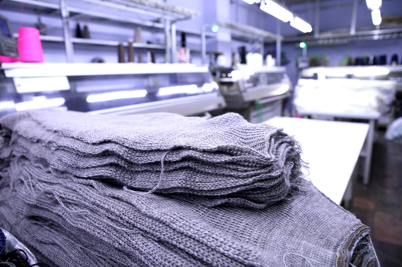 Stos złożonych szarych dzianin na pierwszym planie w hali produkcyjnej, w tle widać maszyny dziewiarskie oraz regały z innymi materiałami.