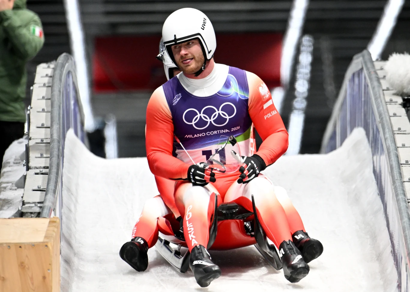 Zjazdowiec na torze saneczkowym w trakcie zimowych igrzysk olimpijskich, ubrany w czerwono-biały kombinezon, z kaskiem ochronnym na głowie oraz widocznym numerem startowym i logotypami olimpijskimi.