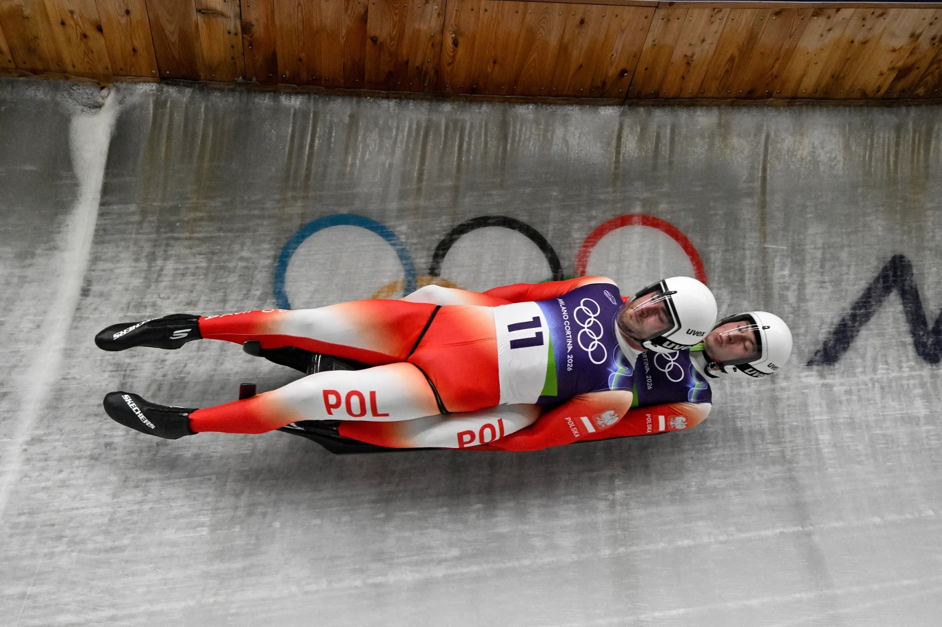 Dwóch polskich zawodników w stroju startowym i kaskach zjeżdża na sankach torowych podczas zawodów olimpijskich, na tle widoczne są obręcze olimpijskie.