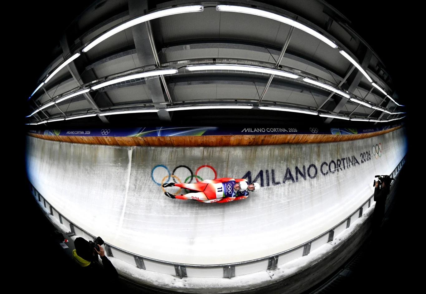 Zawodnik startujący na torze saneczkowym, jadący na sankach obok napisu Milano Cortina 2026 i olimpijskich kół na lodowej ścianie, a całość uchwycona szerokokątnym obiektywem pod zadaszoną trybuną.