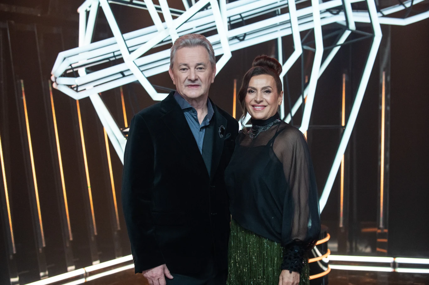 Finaliści z drużyny Andrzeja Piasecznego Dwie elegancko ubrane osoby stoją obok siebie na tle nowoczesnej scenografii z geometrycznymi świecącymi elementami w formie linii.