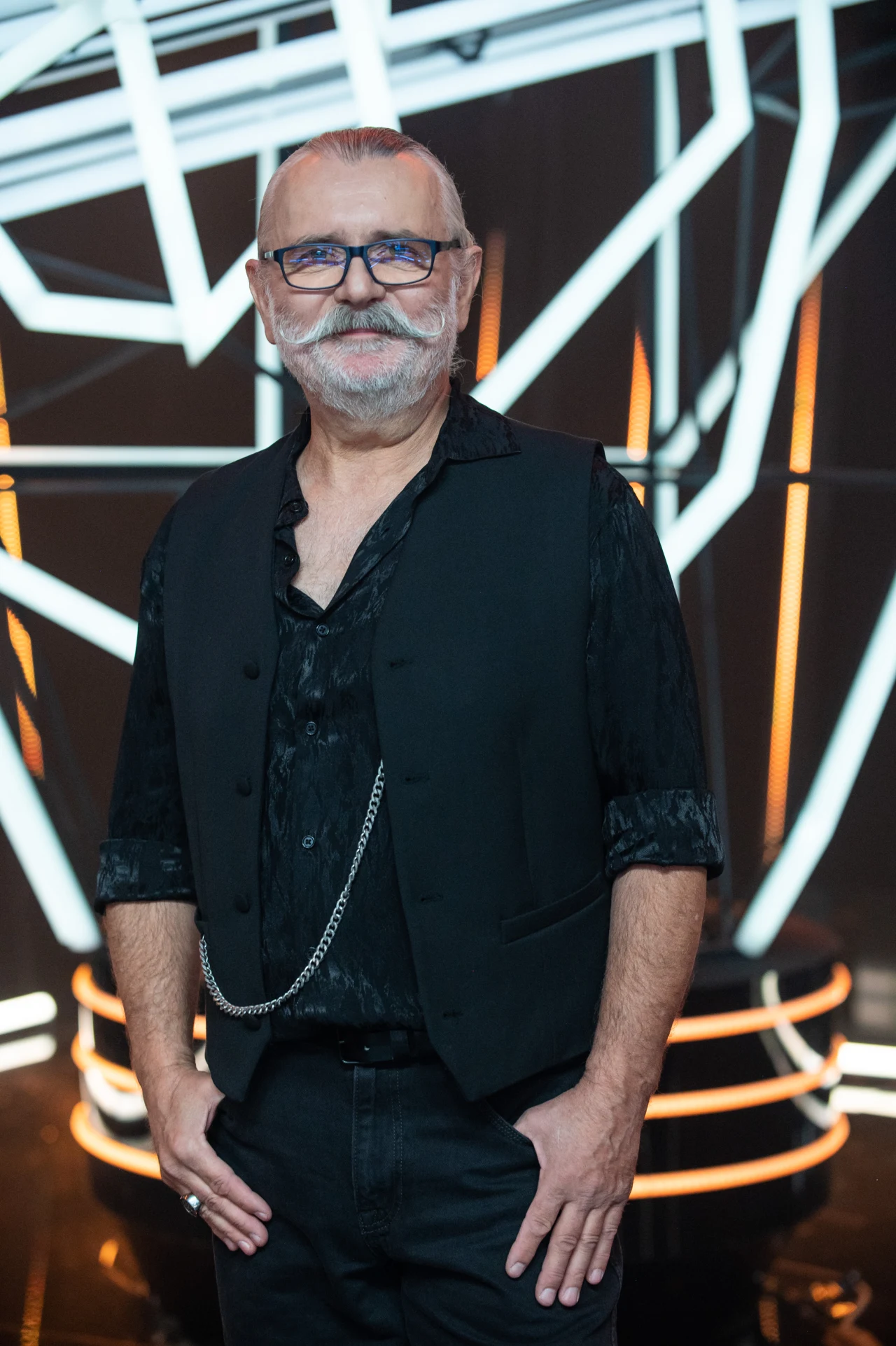 Artur Słowik to finalista "The Voice Senior" z drużyny Majki Jeżowskiej Starszy mężczyzna z siwymi włosami i brodą, ubrany elegancko na czarno, z kamizelką oraz okularami, stoi pewnie w nowoczesnym, abstrakcyjnym otoczeniu z geometrycznymi neonowymi liniami w tle.