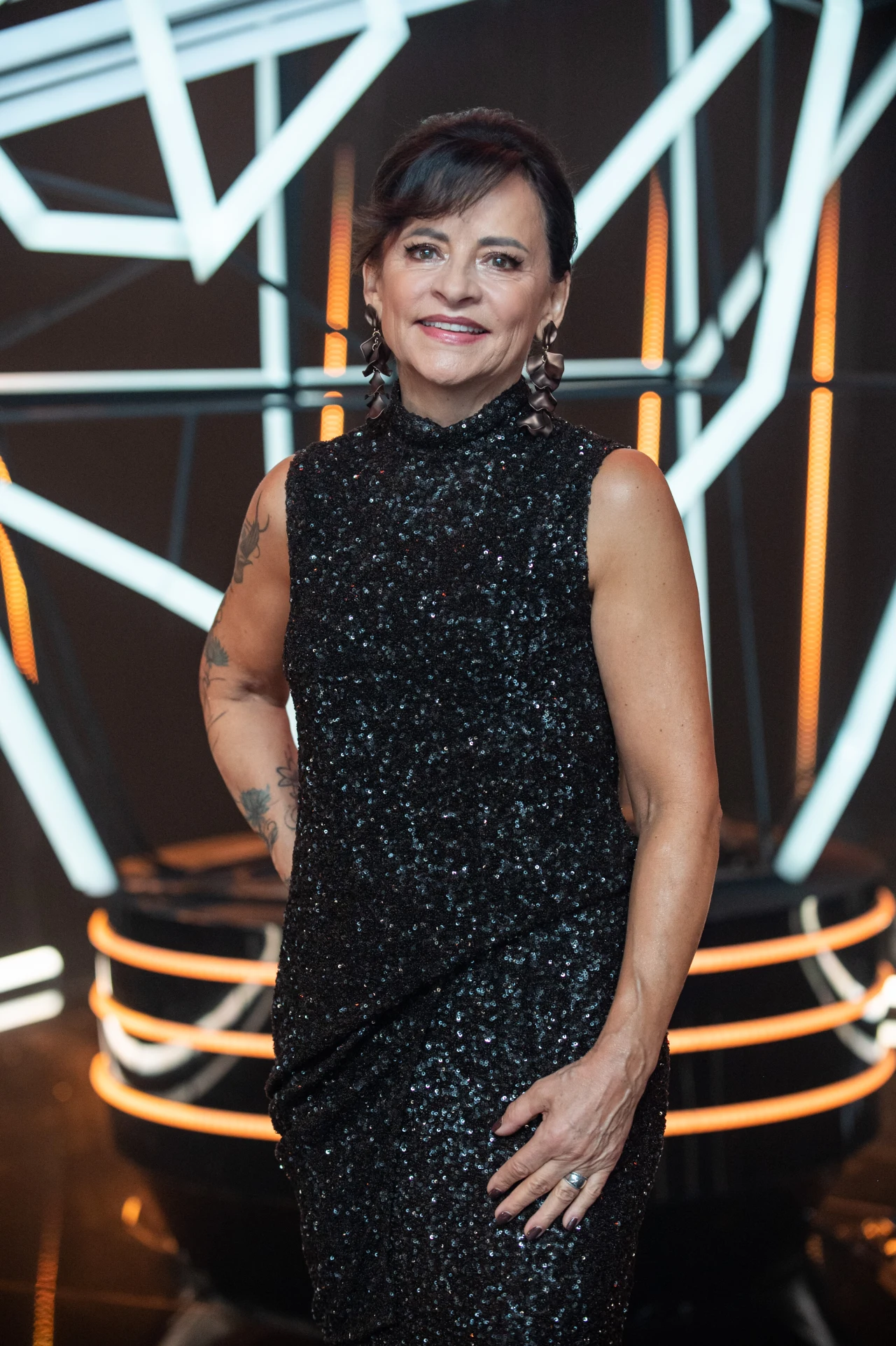 Małgorzata Skrzypczak została finalistką "The Voice Senior" Kobieta w eleganckiej, czarnej, błyszczącej sukience pozuje na tle geometrycznych instalacji świetlnych, uśmiechając się z pewnością siebie.