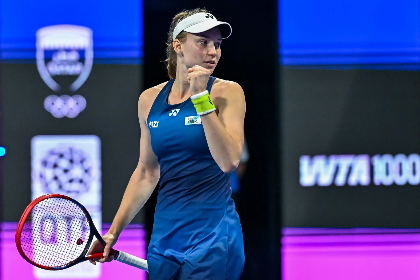 Tenisistka ubrana w niebieski strój sportowy z opaską na głowie trzyma rakietę w ręce i wykonuje gest triumfu, stojąc na korcie, w tle widoczne są tablice wyników i logo turnieju.