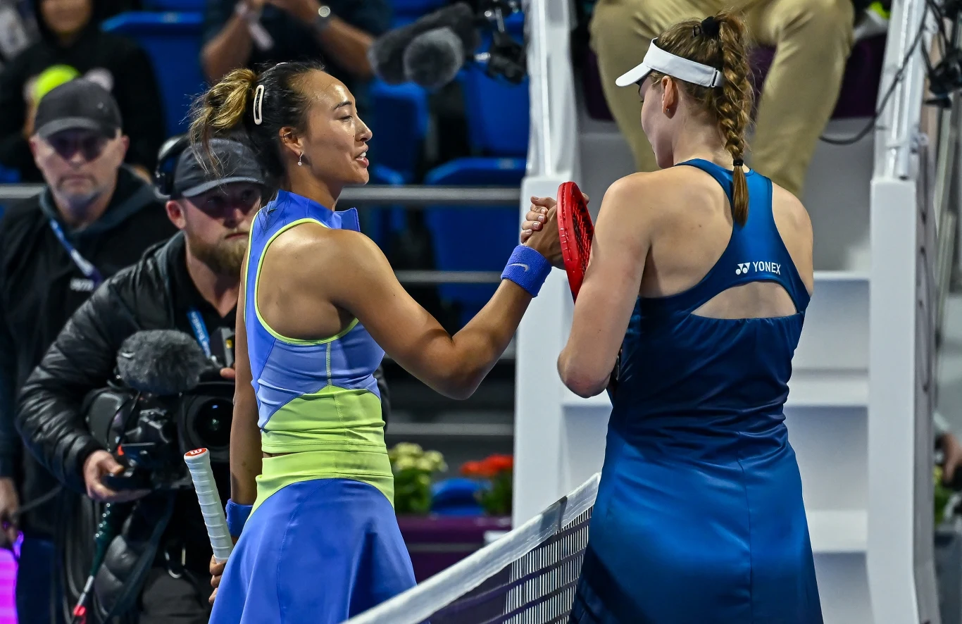 Dwie tenisistki gratulują sobie wzajemnie po zakończonym meczu, stojąc po przeciwnych stronach siatki na korcie, otoczone przez widzów oraz obsługę turnieju.