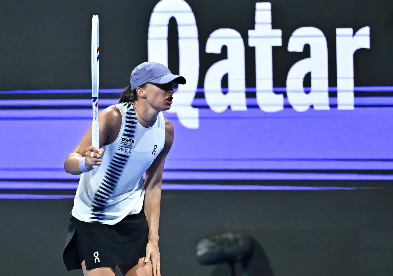 Tenisistka w sportowym stroju i czapce szykuje się do zagrania podczas meczu na tle dużego, podświetlonego napisu 'Qatar'.