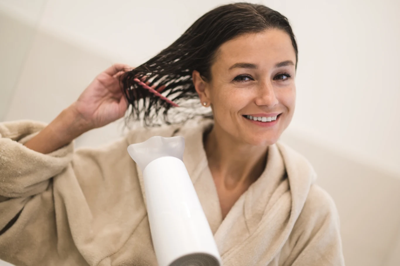 Młoda kobieta w szlafroku suszy mokre włosy suszarką, delikatnie przeczesując je grzebieniem, uśmiecha się do aparatu, w tle jasne, neutralne wnętrze łazienki.
