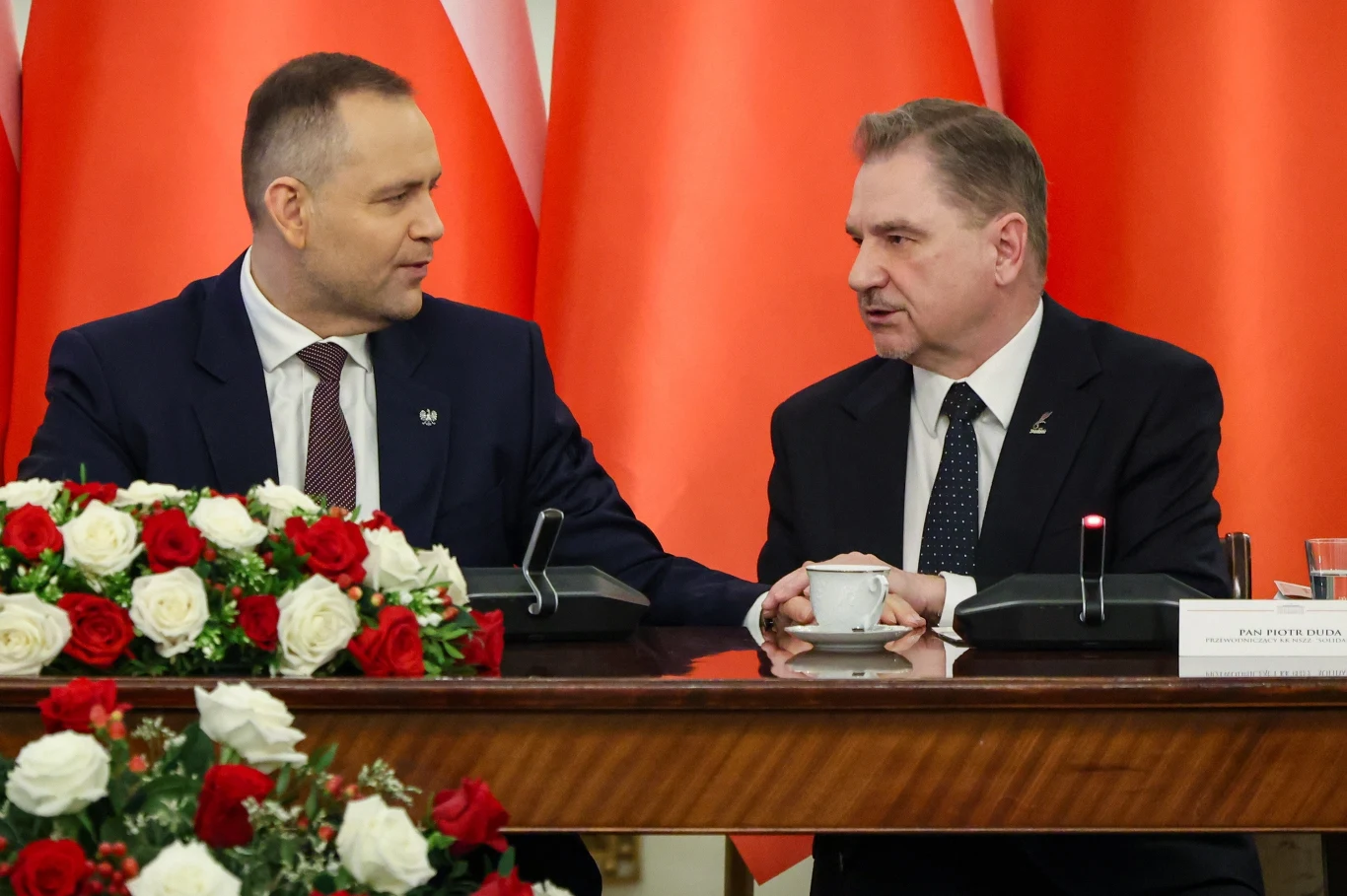 Prezydent RP Karol Nawrocki i przewodniczący NSZZ Solidarność Piotr Duda Dwóch mężczyzn w garniturach rozmawia ze sobą przy stole konferencyjnym, przy którym stoją mikrofony i filiżanka. W tle znajdują się biało-czerwone flagi oraz kompozycje kwiatowe z róż.