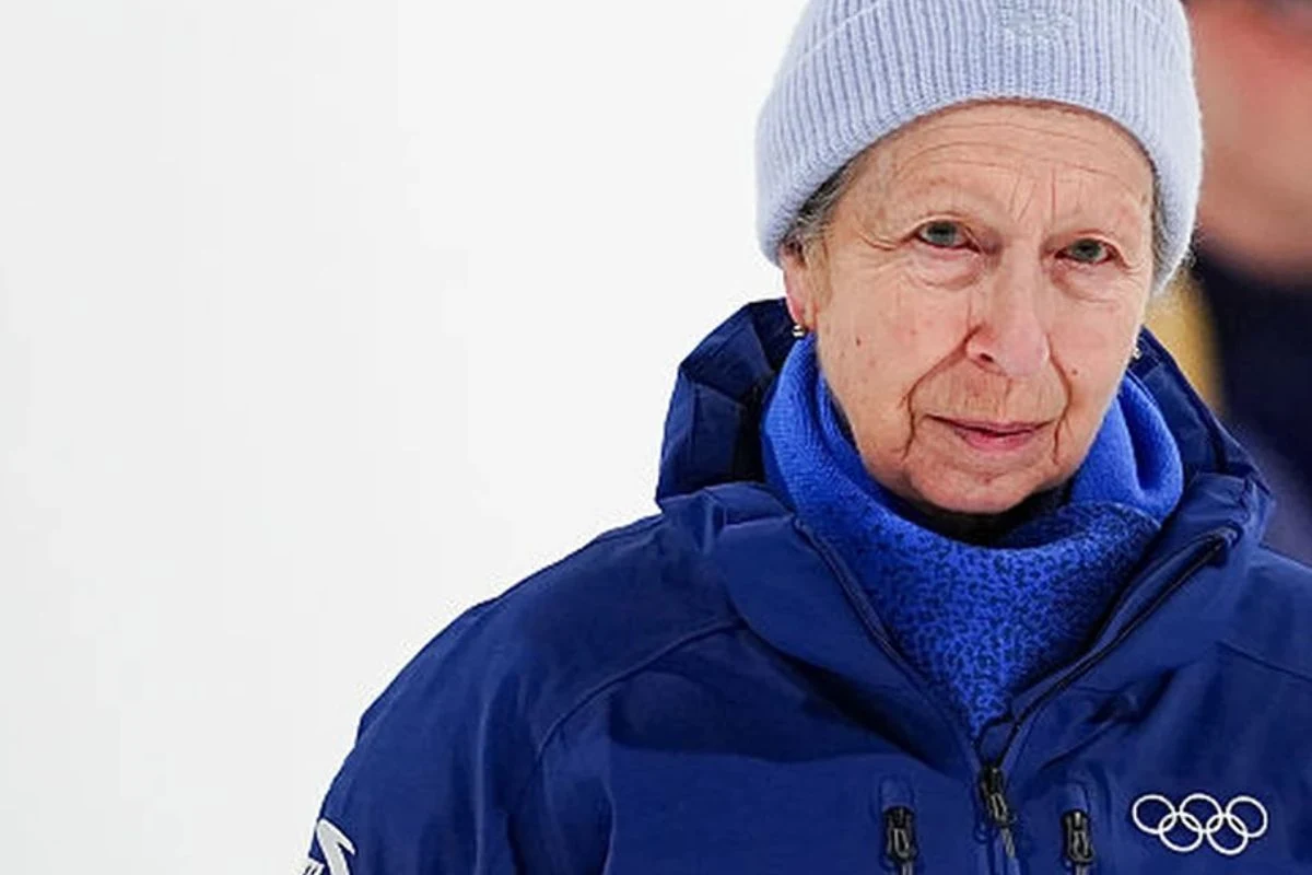 Starsza kobieta w niebieskiej kurtce z logo olimpijskim oraz szarej czapce, patrząca poważnie przed siebie na tle jasnego, rozmytego tła.