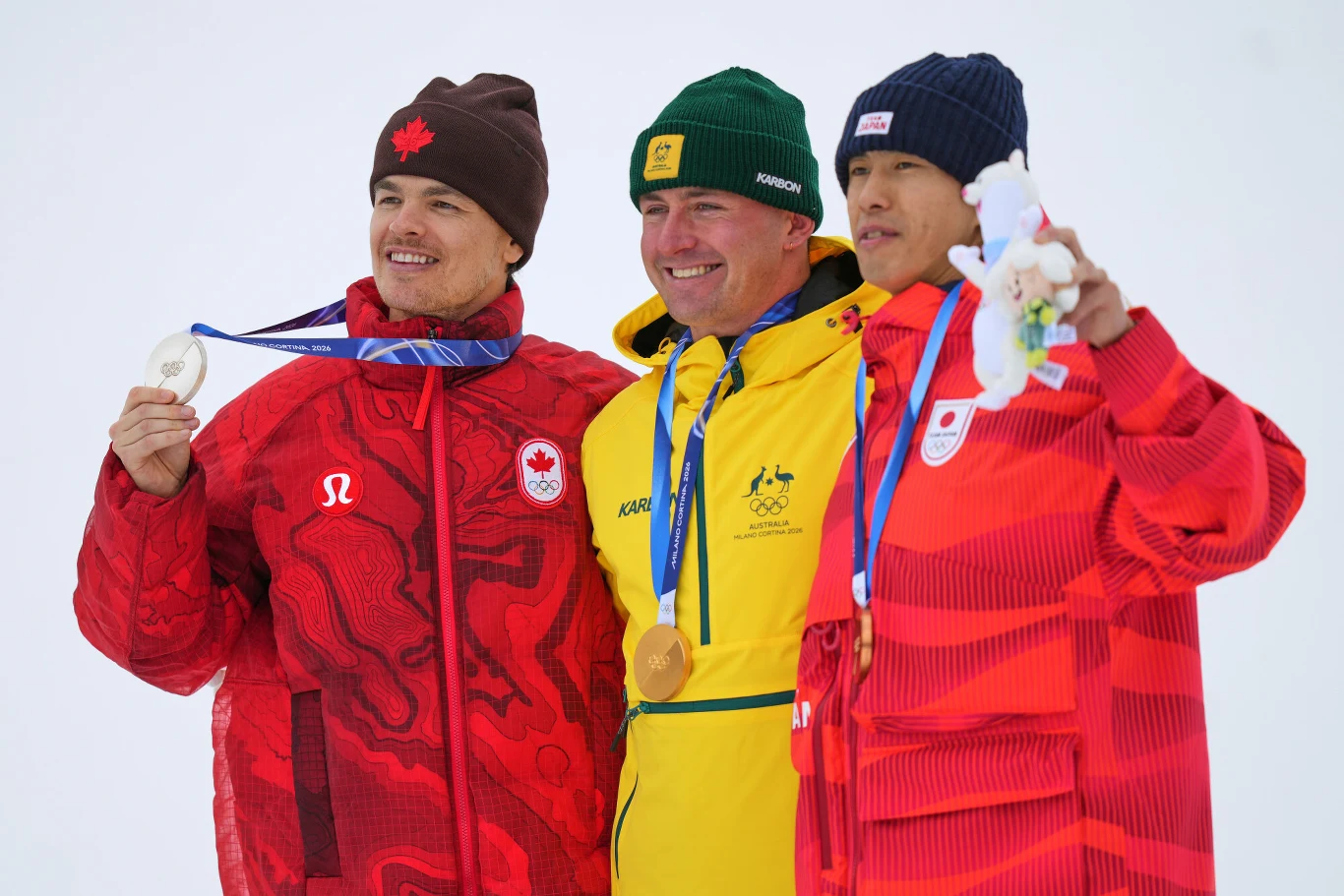Mikael Kingsbury, Cooper Woods i Ikuma Horishima Trzech sportowców w zimowych strojach narodowych stoi razem na podium, prezentując swoje medale olimpijskie i maskotkę. Zwycięzca w centrum w żółtym ubraniu trzyma złoty medal, pozostali zawodnicy mają srebrny i brązowy medal.
