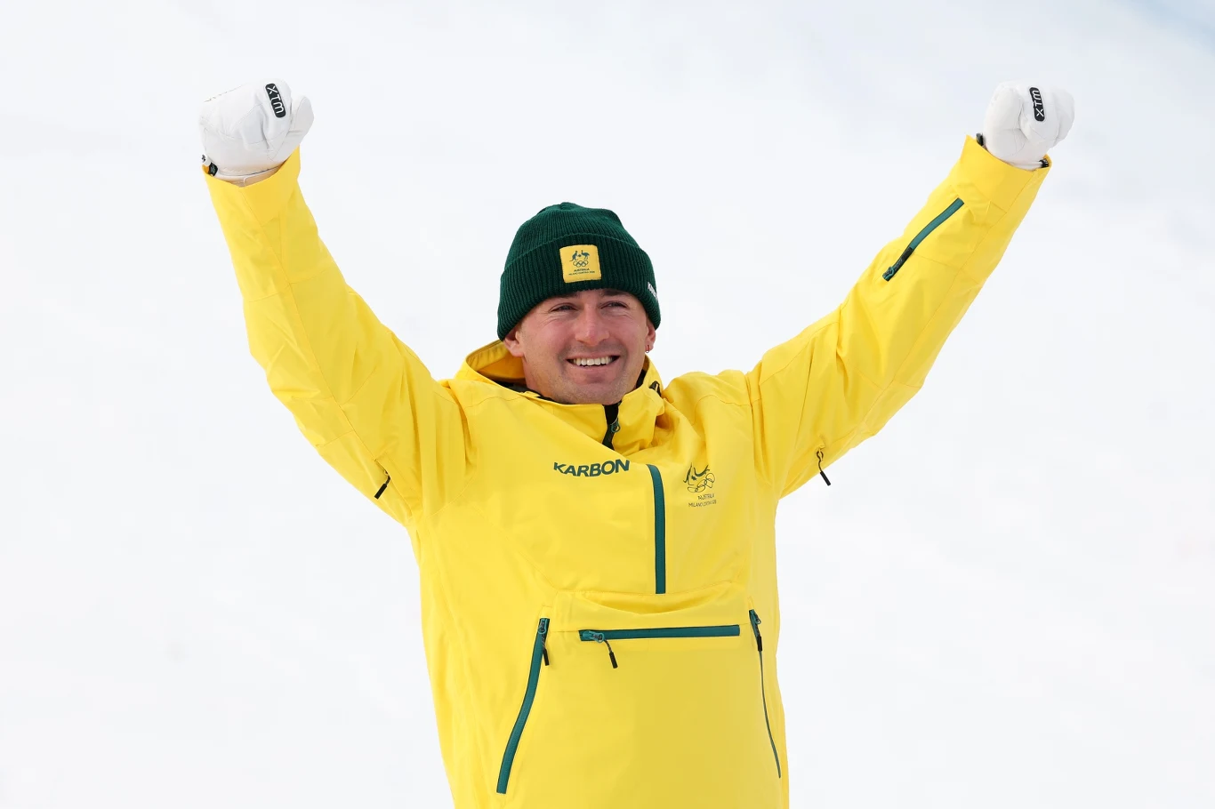 Cooper Woods Mężczyzna w żółtej kurtce sportowej i zielonej czapce stoi z uniesionymi rękami w geście triumfu na tle śniegu, uśmiechnięty, w jasnych rękawiczkach.