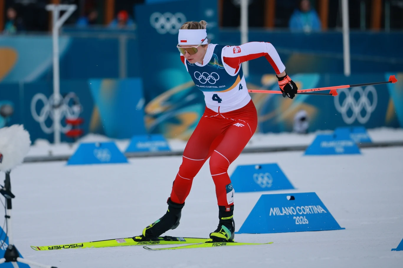 Zawodniczka w biało-czerwonym stroju narciarskim z numerem 4 startuje w biegu narciarskim podczas zimowych igrzysk olimpijskich Milano Cortina 2026, widoczne elementy toru i oznaczenia olimpijskie.
