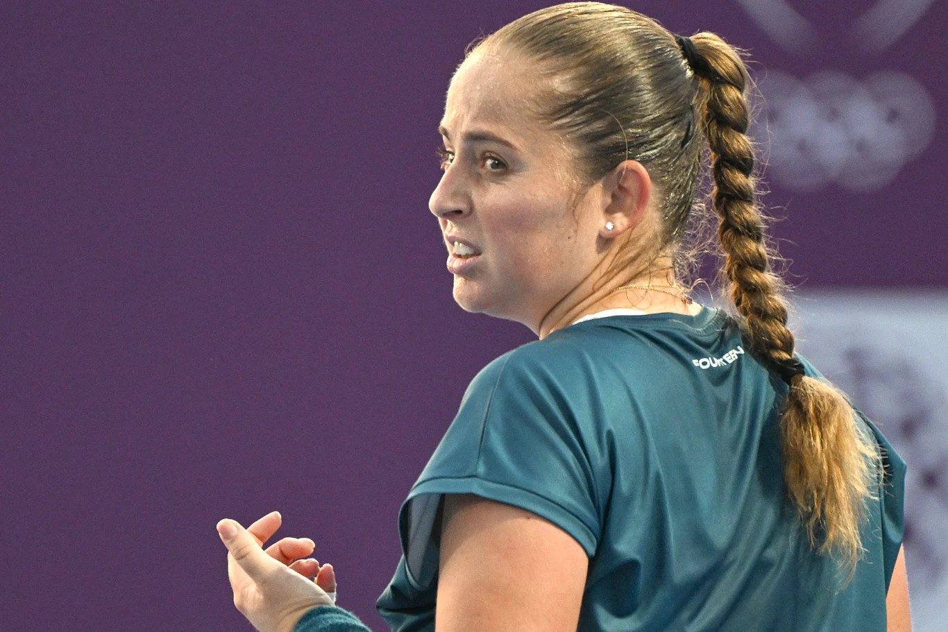 Kobieta o jasnej cerze z długim warkoczem ubrana w sportową koszulkę, odwrócona lekko do tyłu, z wyraźnym wyrazem emocji na twarzy, gestykulująca dłonią na tle fioletowego boiska sportowego.