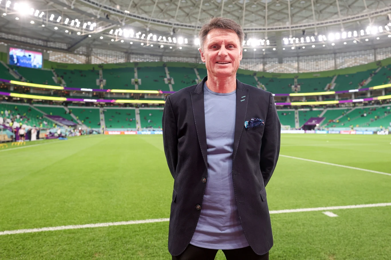 Janusz Michallik Mężczyzna w marynarce i koszulce stoi na murawie stadionu piłkarskiego, w tle trybuny i dach areny sportowej.