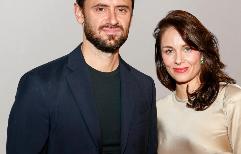 Dwie elegancko ubrane osoby stoją blisko siebie i uśmiechają się, na jasnym tle. Mężczyzna w ciemnej marynarce, kobieta w jasnej, satynowej bluzce z delikatnymi kolczykami.