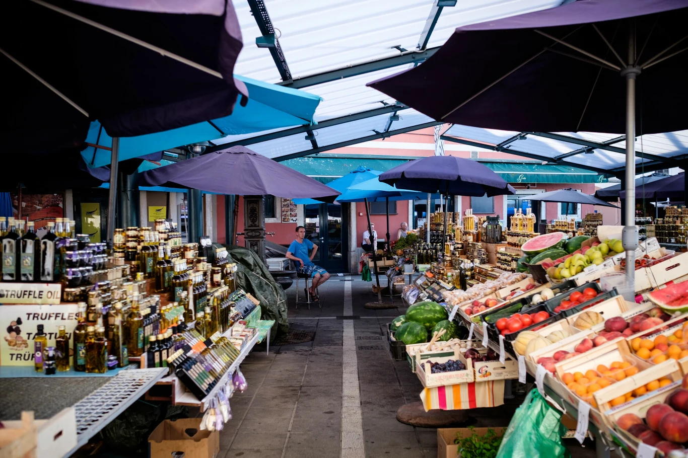 Ceny na bazarach przyprawiają Chorwatów o ból głowy. Kolorowe stoisko na targu z owocami, warzywami oraz butelkami oliwy i innych lokalnych produktów, osłonięte dużymi parasolami. Pośrodku alejka prowadząca w głąb targu, gdzie widoczni są sprzedawcy oraz klienci.