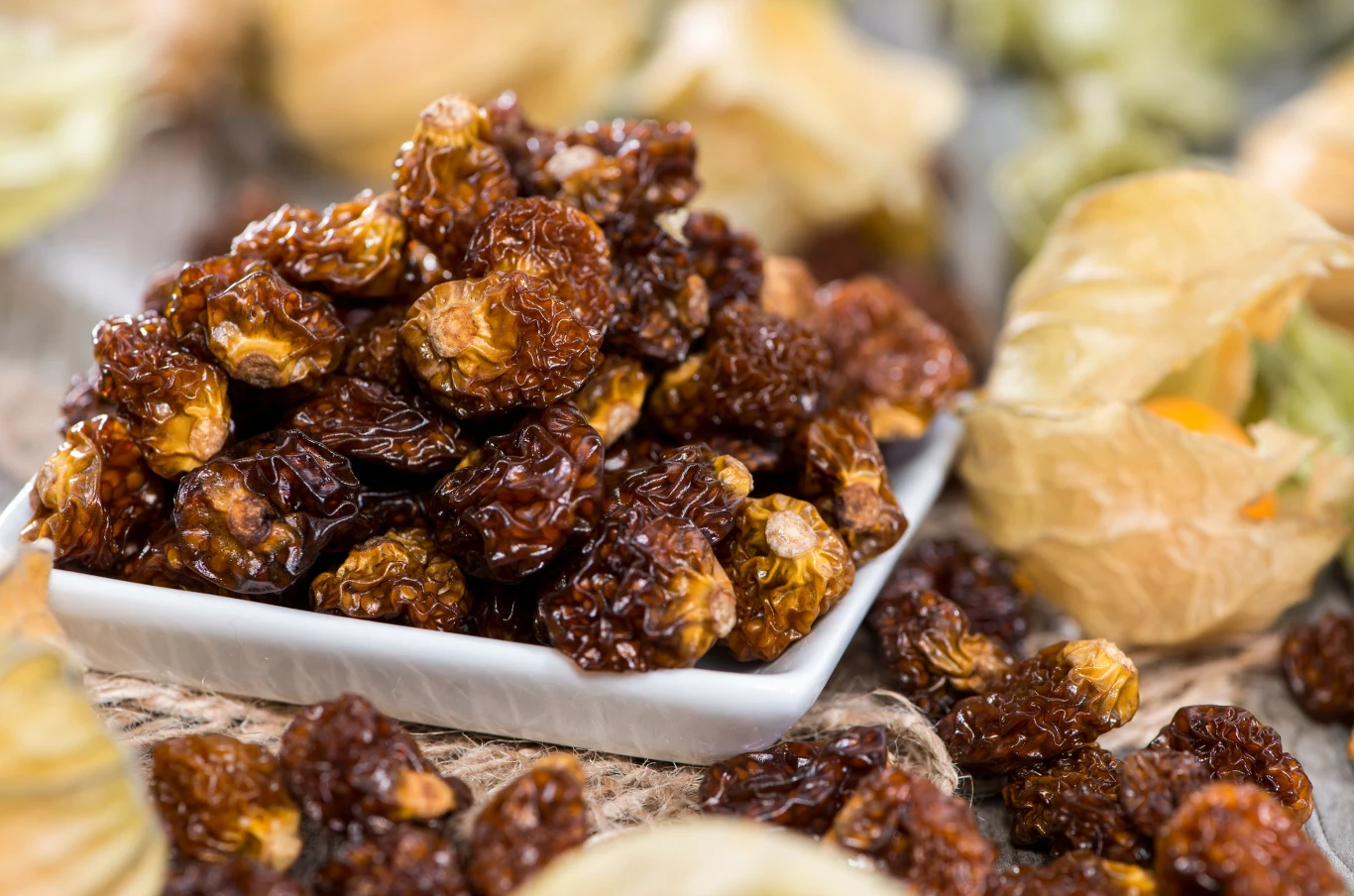 Miechunkę suszoną można jeść przez cały rok. Wystarczy kupić ją w sklepie Stos suszonych owoców miechunki leżący na białej miseczce, wokół której rozłożone są świeże owoce w jasnych osłonkach. Kompozycja skupia się na fakturze i barwie owoców.