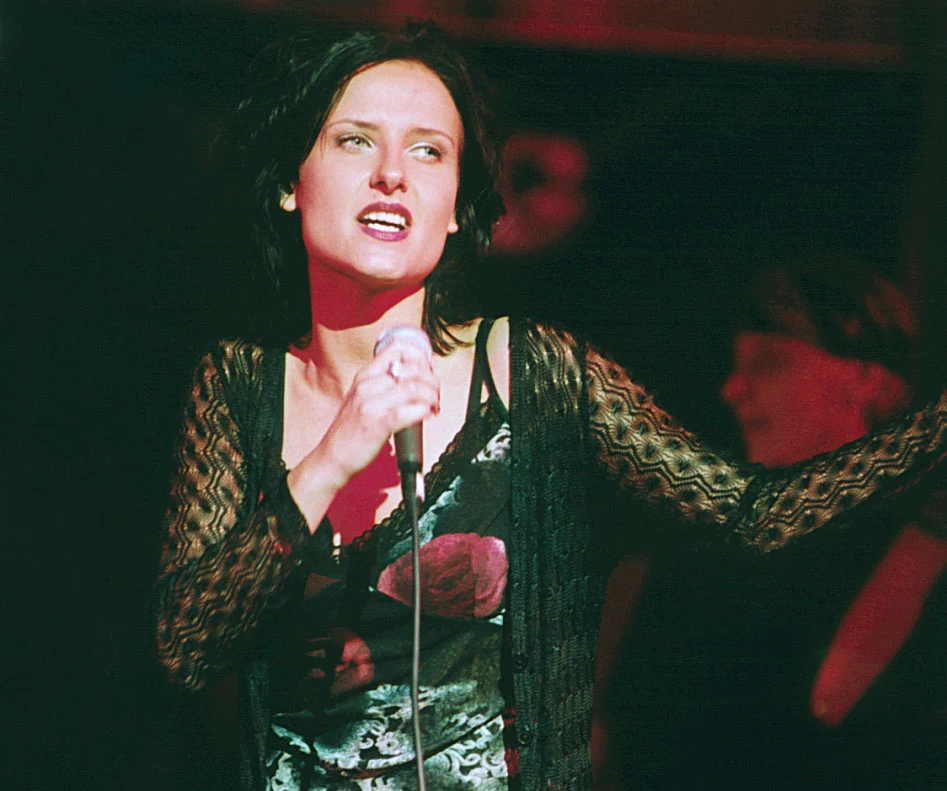 Patrycja Markowska w 2001 r. Kobieta trzyma mikrofon i śpiewa na scenie oświetlonej czerwonym światłem, w tle niewyraźne sylwetki innych osób.