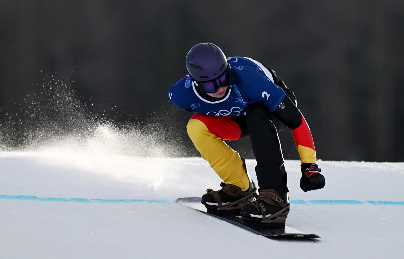 Leon Ulbricht Snowboardzista w specjalistycznym stroju oraz kasku pokonuje ośnieżony stok, pochylony dynamicznie w trakcie jazdy, wokół śnieg rozpryskuje się spod deski.