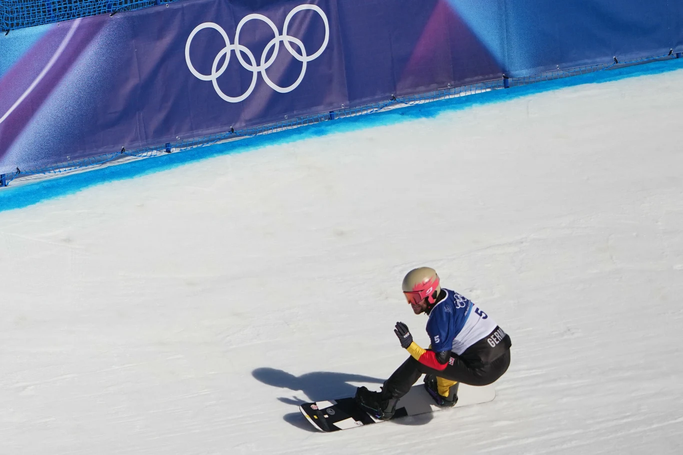 Martin Nörl Zawodnik na snowboardzie w sportowym stroju i kasku zjeżdża po stoku w trakcie zimowych igrzysk olimpijskich, w tle widoczna flaga z logo olimpijskim.