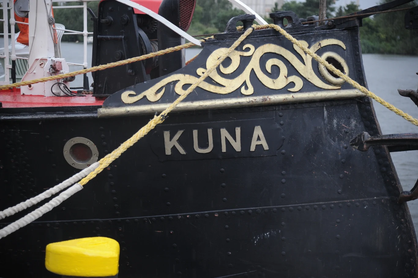Burta czarnej łodzi z widocznymi ozdobnymi złotymi zdobieniami oraz napisem 'KUNA', statek przycumowany do nabrzeża, na pierwszym planie żółty element cumowniczy.