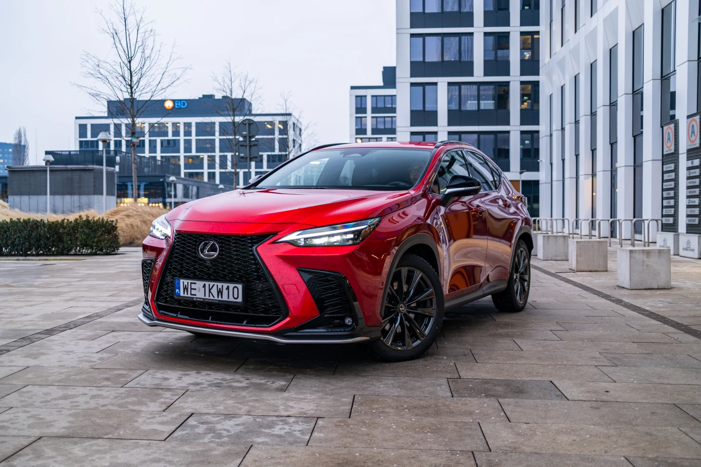 Czerwony samochód marki Lexus NX zaparkowany na tle nowoczesnych budynków biurowych, otoczenie miejskie, brukowana nawierzchnia, niewielka ilość roślinności.
