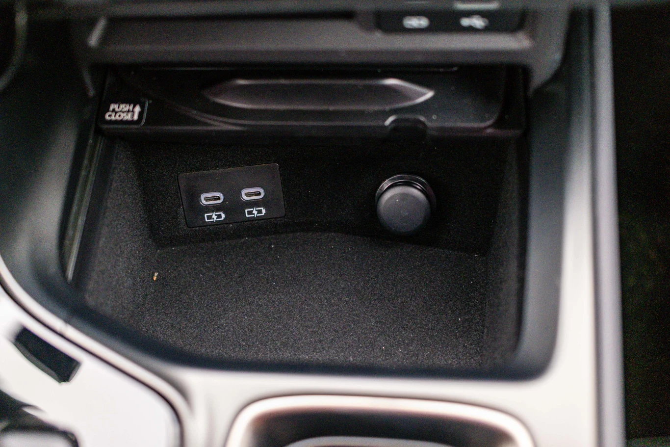 Panel środkowy samochodu z dwoma gniazdami USB oraz gniazdem zasilania 12V, umieszczonymi w schowku wyłożonym czarnym materiałem.