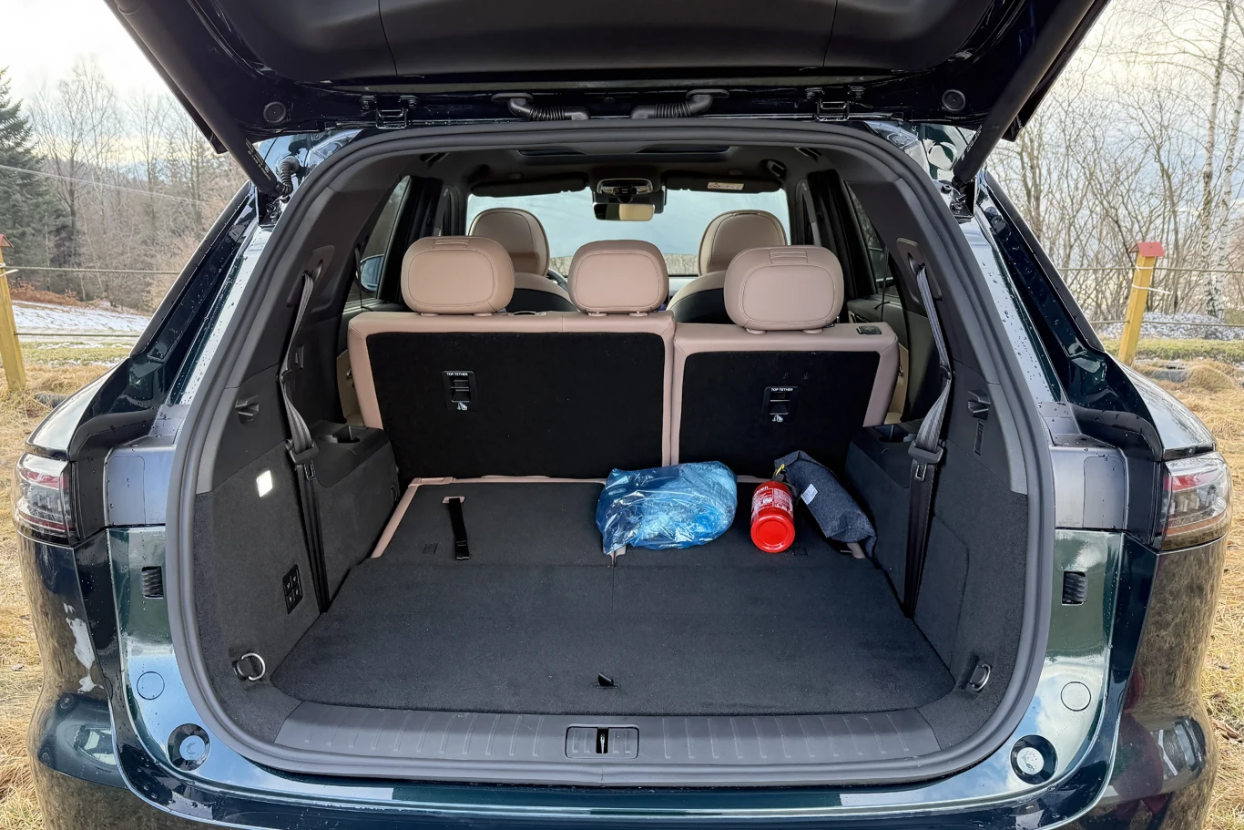 Otwarty bagażnik samochodu terenowego z trzema siedzeniami w drugim rzędzie, w środku widoczna apteczka, butelka oraz złożona kurtka. W tle widać fragment krajobrazu z drzewami i trawą.