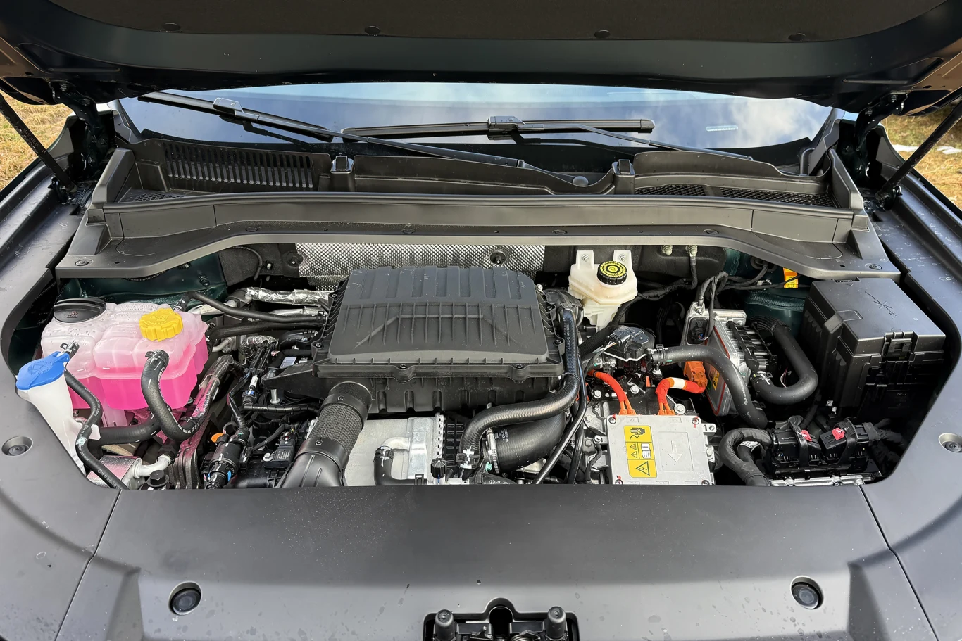 Komora silnika samochodowego z dobrze widocznymi przewodami, zbiornikami płynów oraz osłoną silnika, elementy w różnych kolorach wskazujące na miejsca serwisowe i instalację elektryczną.