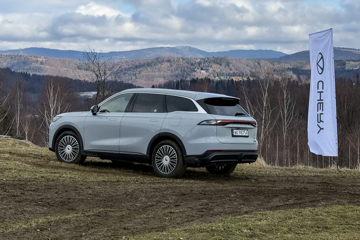 Srebrny samochód terenowy marki Chery zaparkowany na lekko podmokłej łące, z widokiem na górzysty krajobraz w tle, po prawej stronie widoczny biały baner z logo Chery.