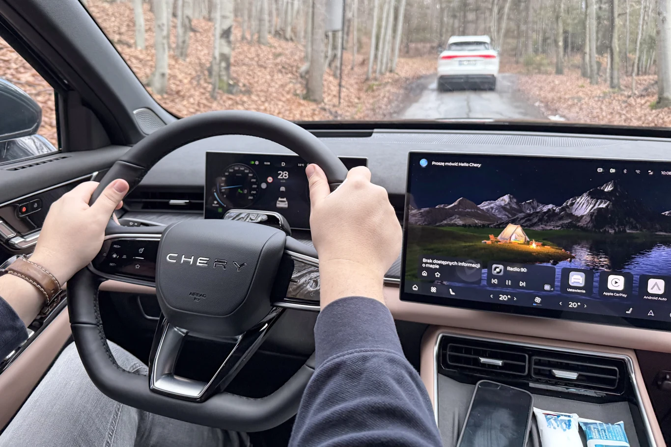 Kierowca prowadzi nowoczesny samochód marki Chery, widoczny kokpit z zaawansowanymi wyświetlaczami cyfrowymi, przed pojazdem leśna droga z jadącym przed nim samochodem, drzewa pozbawione liści, na siedzeniu widoczny telefon i opakowanie chusteczek.