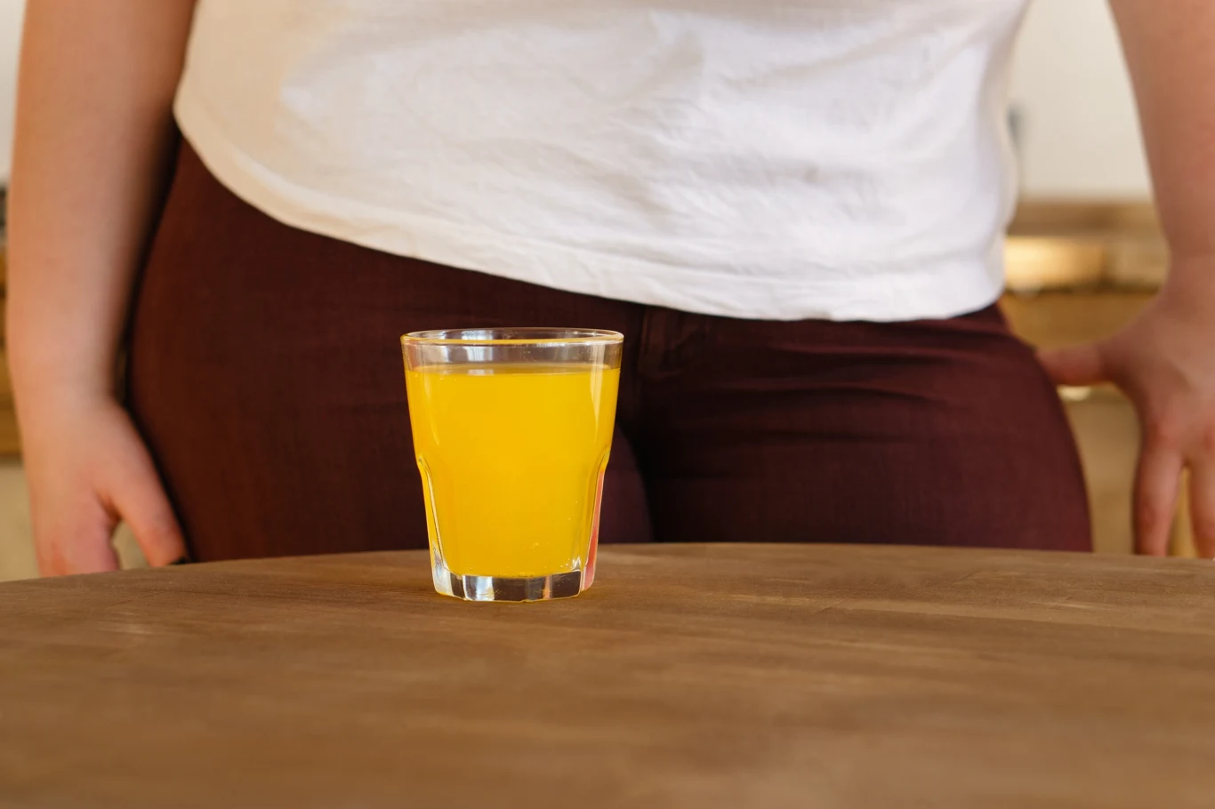 Kolorowe napoje są plagą dzisiejszych czasów. Wiele z nich zawiera toksyczne słodziki Szklanka z żółtym napojem stoi na drewnianym stole, w tle fragment sylwetki osoby w białej koszulce i bordowych spodniach, której ręce opierają się o stół.