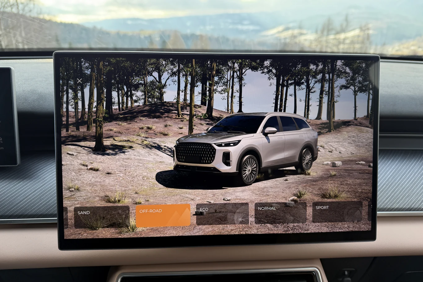 Wyświetlacz pokładowy samochodu prezentuje wizualizację srebrnego SUV-a na tle leśnego terenu. Na ekranie widoczne są opcje trybów jazdy, z wyróżnioną opcją off-road, otoczenie stanowią drzewa i pagórkowaty krajobraz.
