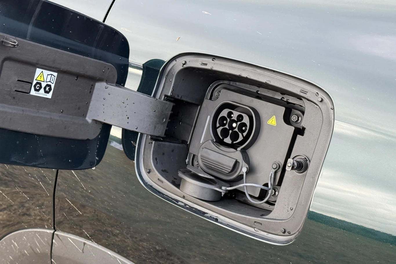 Gniazdo ładowania samochodu elektrycznego odsłonięte po otwarciu klapki, widoczne symbole ostrzegawcze oraz zabezpieczenia, powierzchnia lakieru auta odbijająca otoczenie.