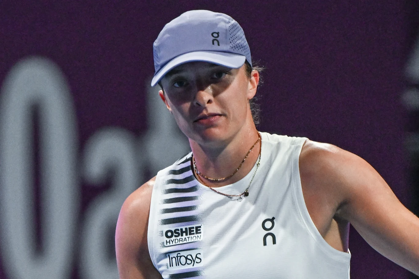 Iga Świątek walczyła o półfinał WTA 1000 w Dosze Młoda kobieta w sportowym stroju i czapce z daszkiem znajduje się na tle rozmytego napisu, nosi białą koszulkę z logo marek sportowych oraz medaliki na szyi, patrzy skierowana w stronę obiektywu.