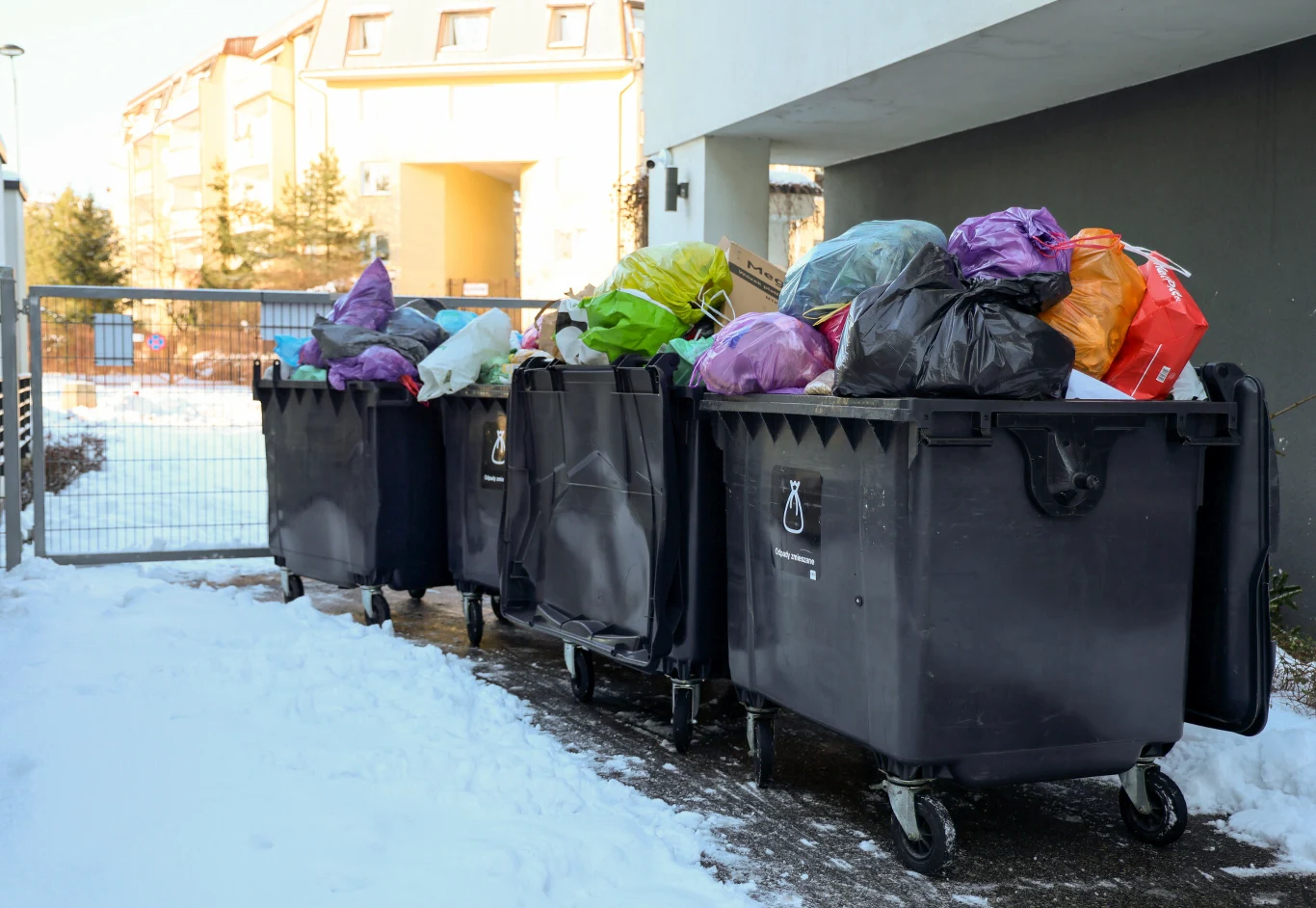 Nieodpowiednia segregacja odpadów może słono kosztować Rzędy dużych pojemników na śmieci ustawionych na zewnątrz obok budynku, przepełnione workami na odpady różnych kolorów, wokół śnieg, w tle ogrodzenie i fragmenty osiedla mieszkaniowego.