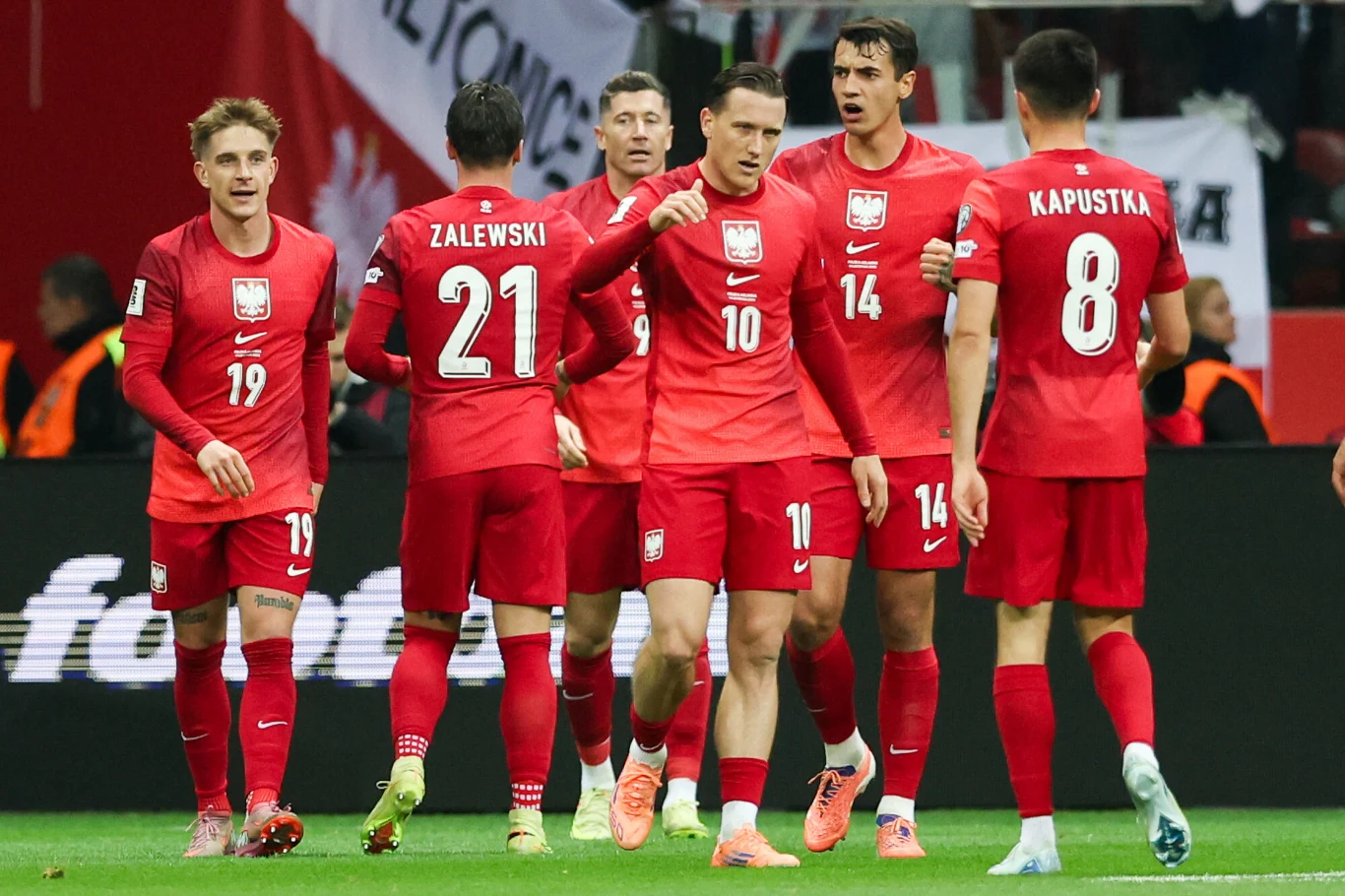 Grupa piłkarzy reprezentacji Polski w czerwonych strojach świętuje na murawie po zdobyciu bramki. W tle widoczni kibice oraz fragmenty trybun.