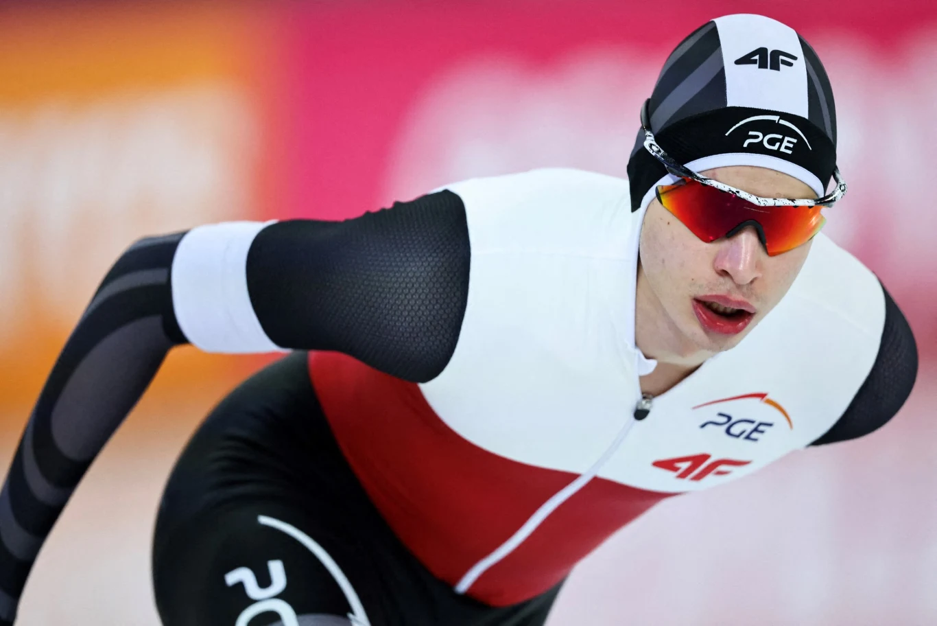 Zawodnik łyżwiarstwa szybkiego w profesjonalnym stroju startowym, z logo sponsorów, pochylony w dynamicznej pozycji podczas wyścigu na lodowisku