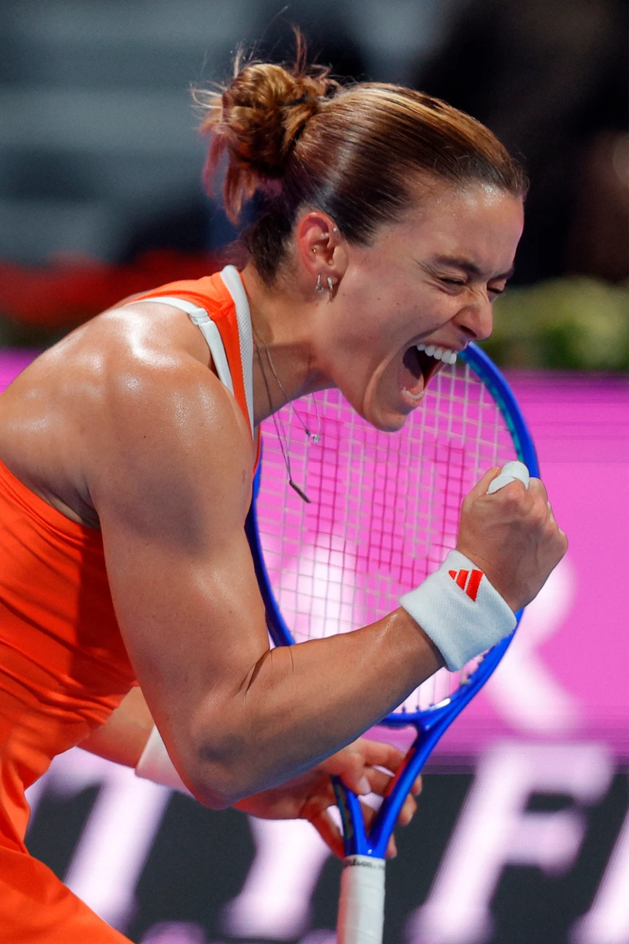 Maria Sakkari Sportsmenka w pomarańczowym stroju z zaciśniętą pięścią i otwartymi ustami wyraża silne emocje zwycięstwa podczas meczu tenisowego, trzymając rakietę w lewej ręce.